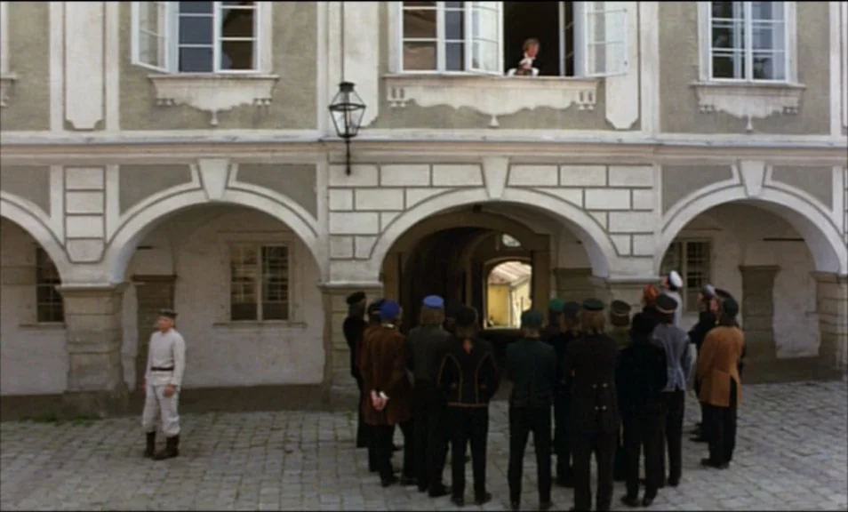 Woyzeck (1979) movie still: wide — A stone courtyard features an arched facade with a man watching from a window. A group of men stands together in…