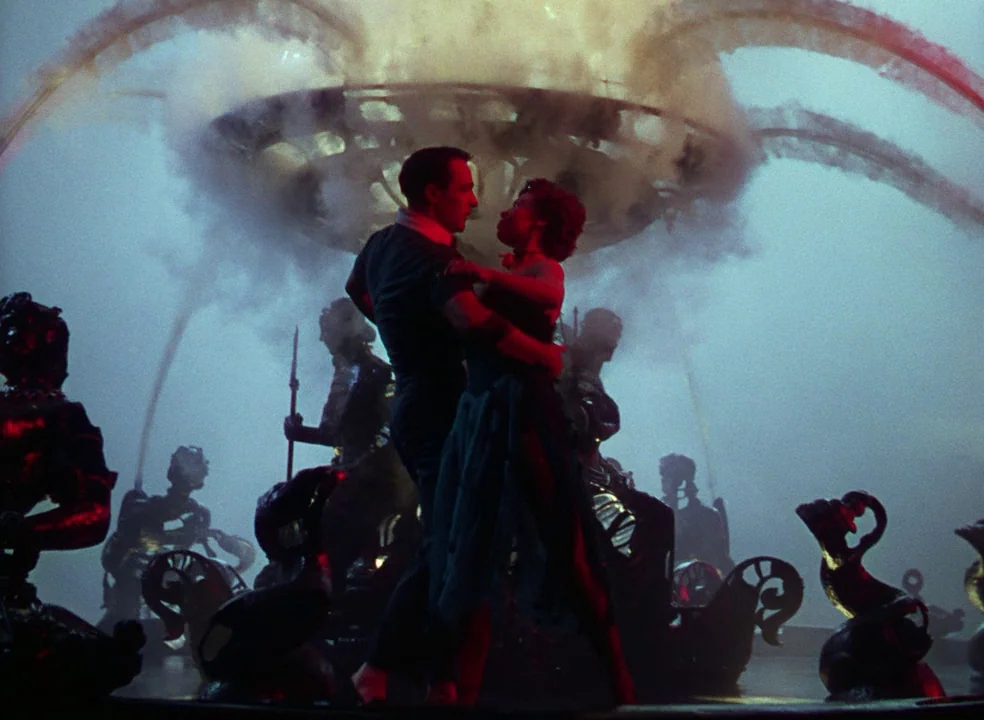 An American In Paris (1951) movie still: fullbody — A dark outdoor space features a large ornamental fountain spraying mist into the air. A man and…