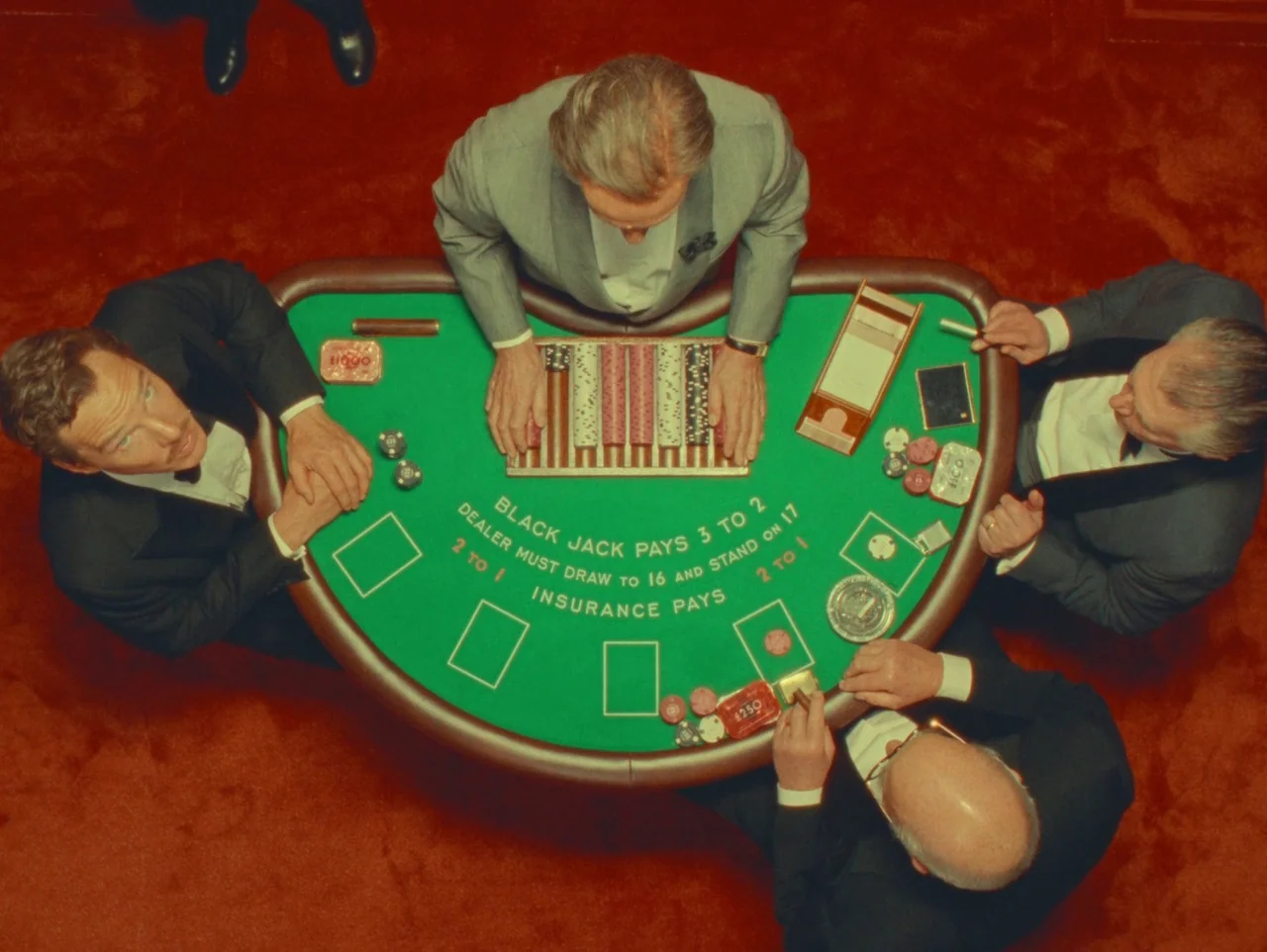 The Wonderful Story of Henry Sugar (2023) movie still: wide — Four men in formal suits sit around a vibrant green blackjack table. They are placing…