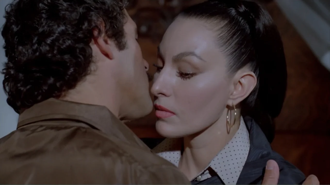 Toña Machetes (1985) movie still: closeup — An indoor space features dark wood panelling in the background behind two close subjects. A woman and a…