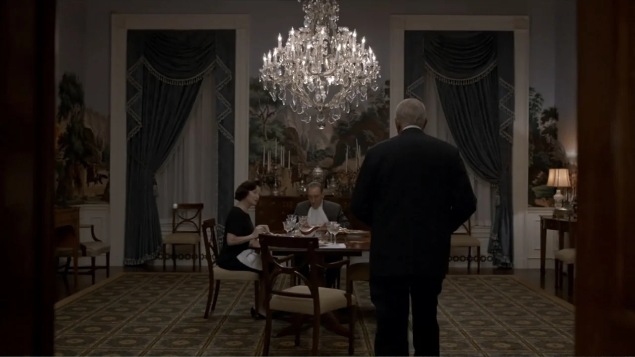 All the Way (2016) movie still: fullbody — A formal dining room featuring a large crystal chandelier, patterned wallpaper, and blue curtains. Three…