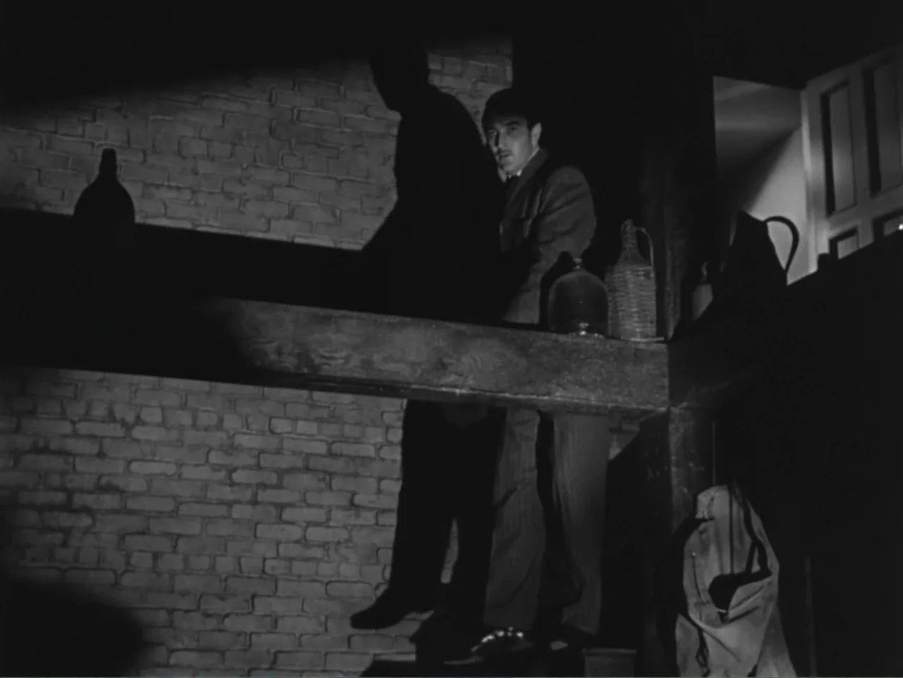 House by the River (1950) movie still: medium — An interior space features an exposed brick wall and a heavy horizontal wooden beam. A man in a suit…
