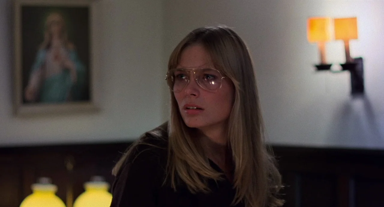 God Told Me To (1976) movie still: closeup — A dimly lit room features a painting of a figure on the wall. A young woman with large glasses glances…