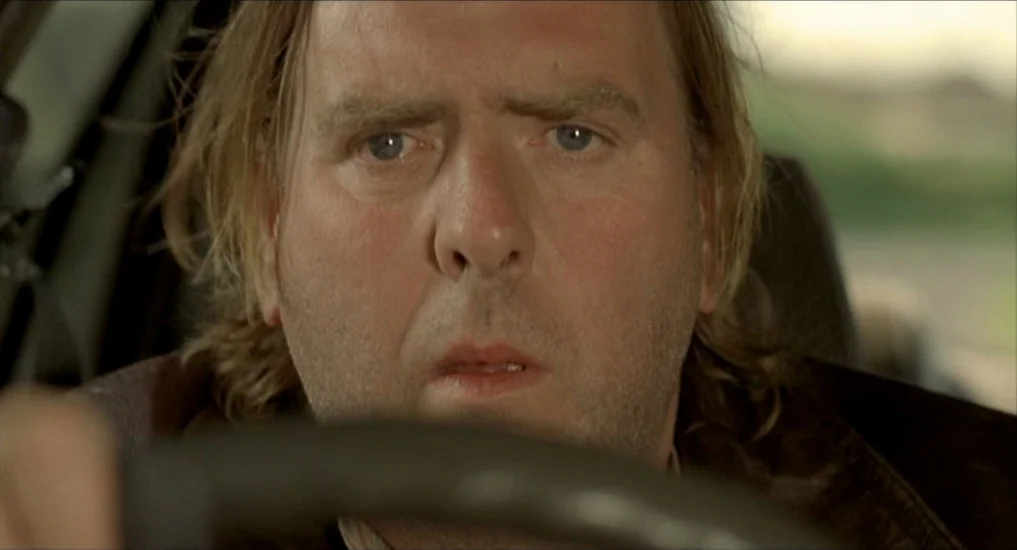 All or Nothing (2002) movie still: closeup — A man sits in the driver seat of a vehicle during the day. The man gazes forward while his hands grip…