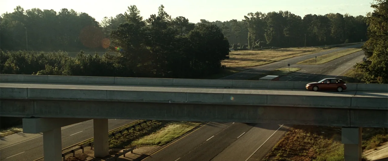 Due Date (2010) movie still: wide — An elevated concrete highway overpass traverses a rural landscape surrounded by dense green forest. A small red…