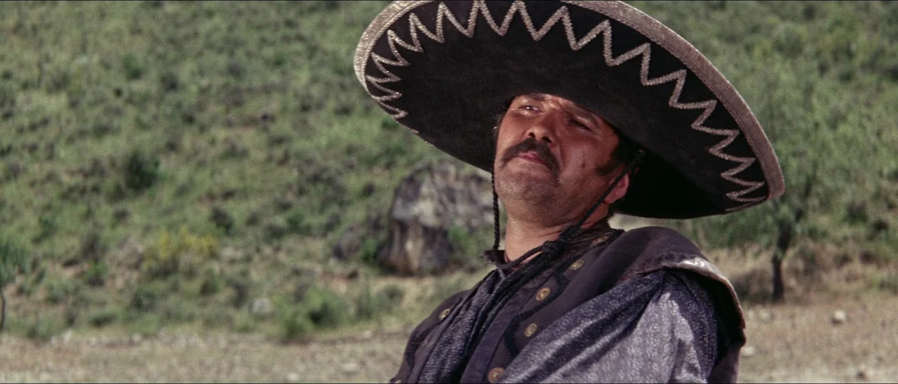 Texas, Adios (1966) movie still: medium — A rural outdoor landscape with scrubby vegetation and a rocky hillside. A man wearing a wide-brimmed…