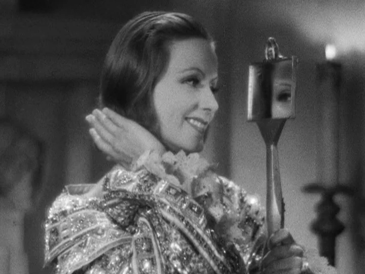 Queen Christina (1933) movie still: closeup — A woman in period-style attire examines her reflection in a small hand mirror. She is indoors, with a…