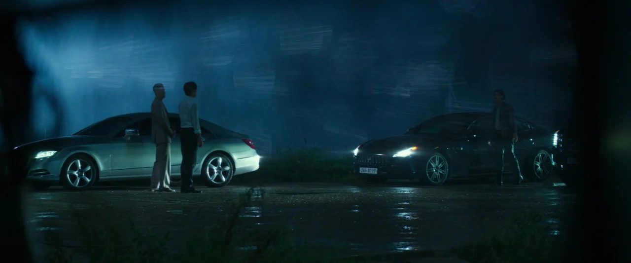 Believer (2018) movie still: wide — Three men stand on a dark, wet asphalt patch between two high-end luxury sedans. The scene is illuminated by the…