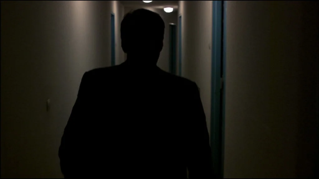 Caché (Hidden) (2005) movie still: medium — A dark hotel corridor features several closed doors along white walls with blue trim. A man in a dark…