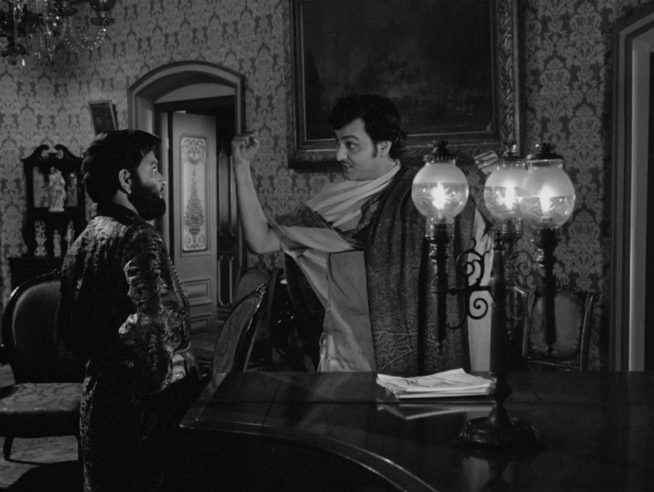 Charulata (1964) movie still: medium — A dimly lit room features ornate floral wallpaper and a large oil painting. Two men stand near a prominent…