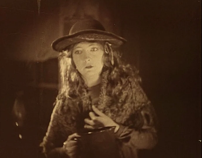 Broken Blossoms (1919) movie still: medium — A dimly lit, rustic interior setting with textured walls and deep shadows. A young woman in a hat holds…