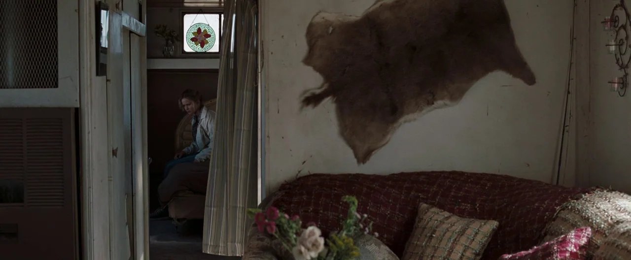 The Burning Plain (2008) movie still: wide — A rustic mobile home interior features a fur pelt mounted on the beige wall. A woman sits on a bed…