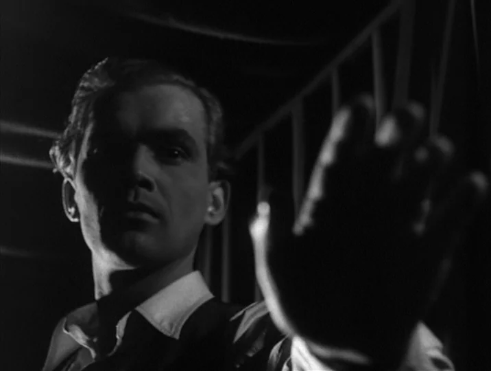 Thirst (1949) movie still: closeup — A dark, cramped interior with metal railing segments visible in the background. A man holds his hand out to…