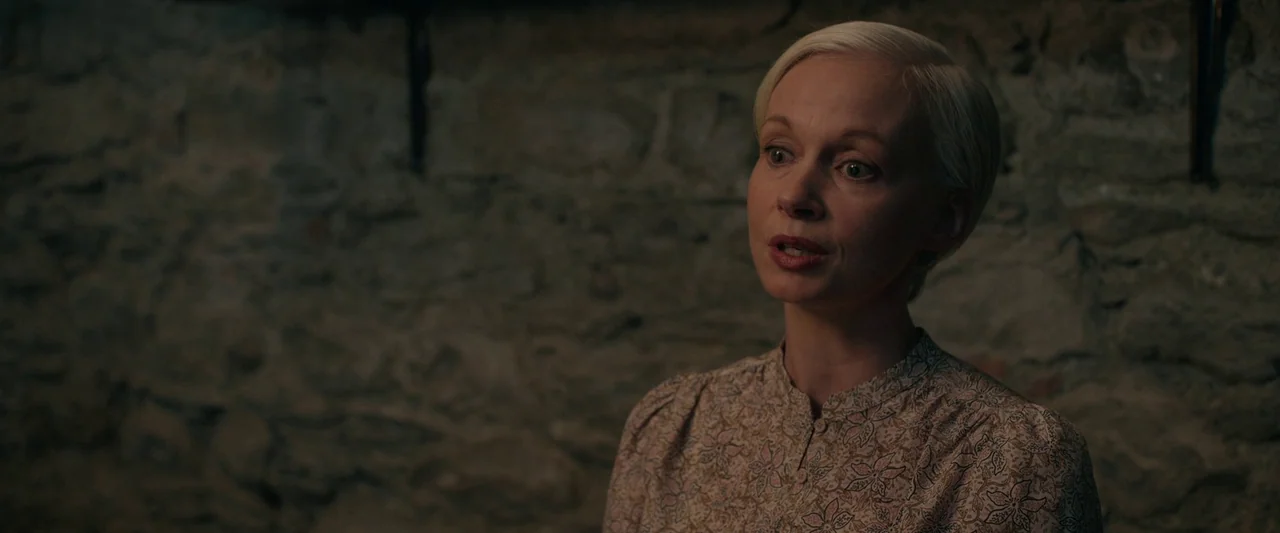 Oddity (2024) movie still: medium — A woman stands in front of a dark, rough-hewn stone wall. She is speaking while looking towards the side of the…