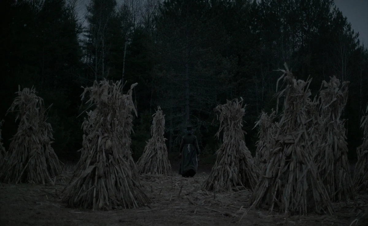 The Witch (2015) movie still: wide — A field filled with bundled, dry corn stalks stands before a dark forest line. A woman in period clothing walks…