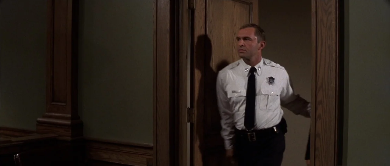 Legally Blonde (2001) movie still: medium — A courtroom interior featuring dark wood paneled walls and a matching wooden door. A uniformed security…