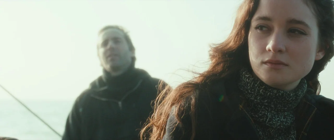 Ginger & Rosa (2012) movie still: medium — A woman with long wavy hair sits on a boat deck in bright daylight. A man stands blurred in the…