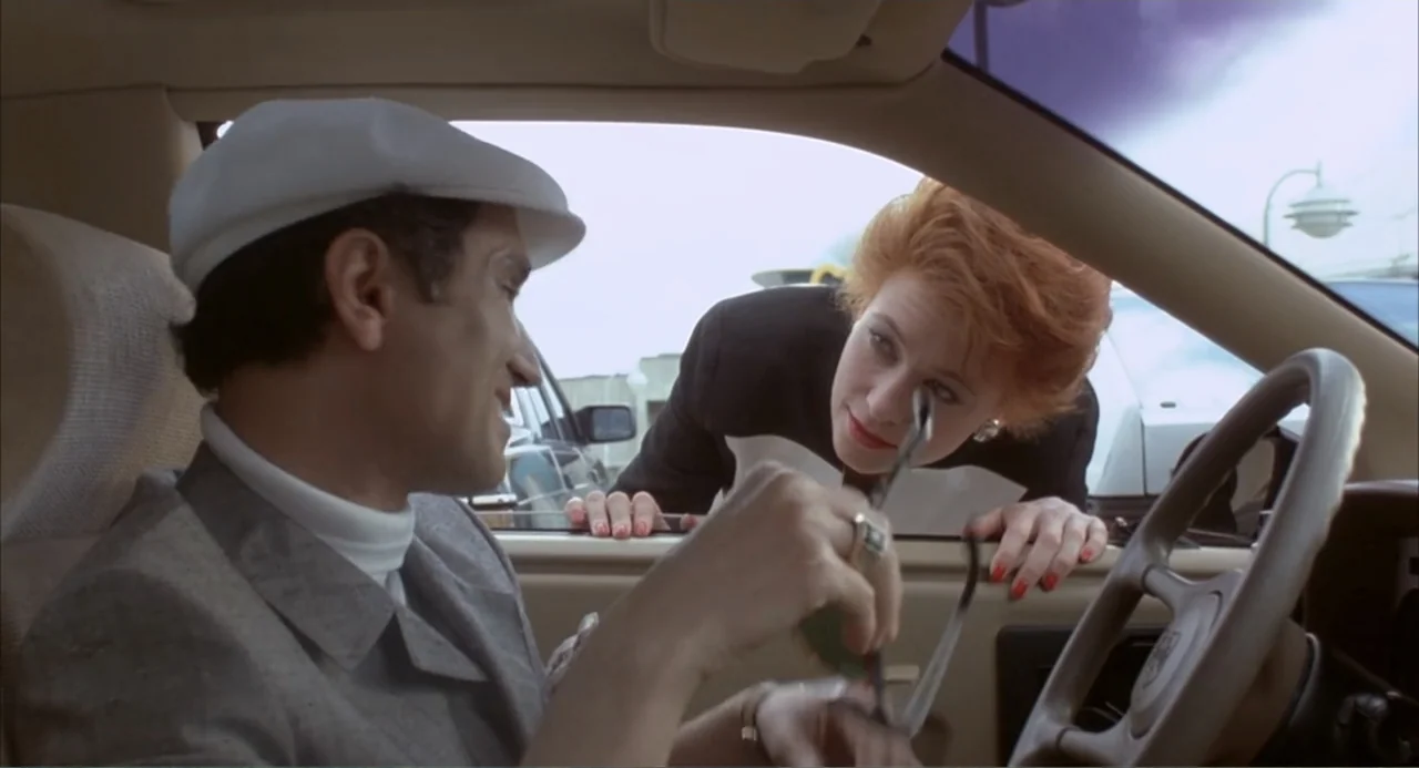 Making Mr. Right (1987) movie still: medium — A man sits in the driver's seat of a parked car in a bright outdoor lot. A woman leans through the…