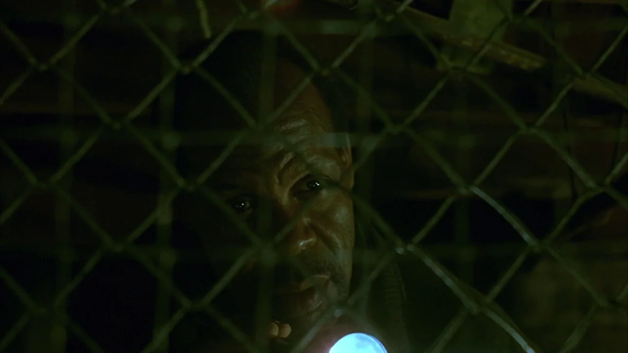 Saw movie still: closeup — A man is partially obscured behind a metal chain link fence in a dark space. He peers through the links while a blue…