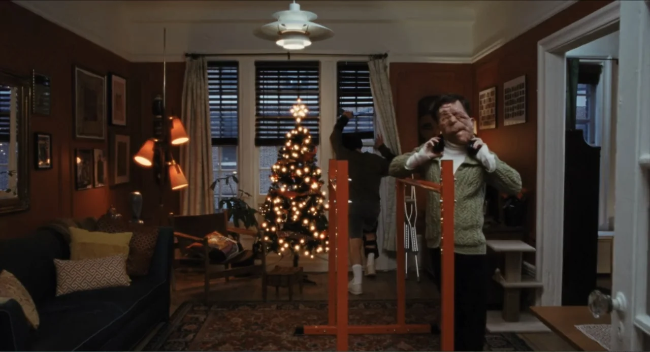 A Different Man (2024) movie still: medium — A furnished living room features dark wood walls, a lit Christmas tree, and framed art. Two men occupy…