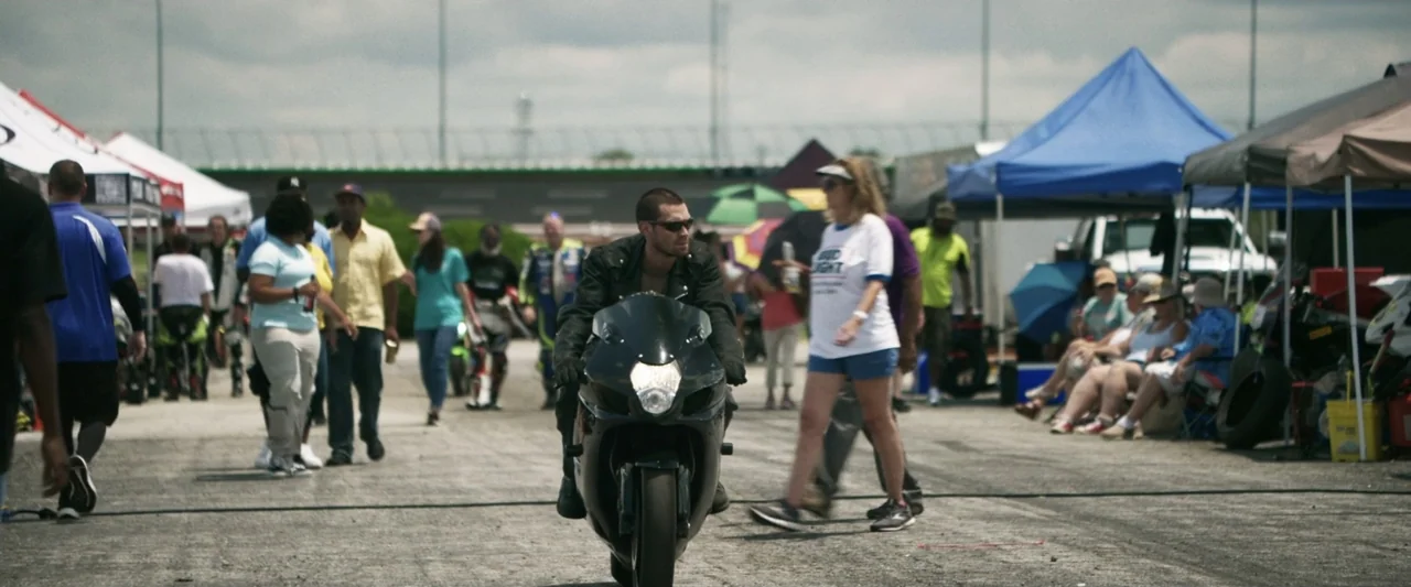 One Fast Move (2024) movie still: medium — A crowded asphalt paddock area is filled with temporary blue tents and groups of people. A man in a black…