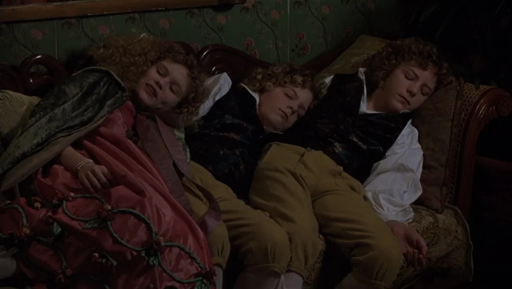 Interview With The Vampire movie still: medium — Three children in period attire sleep side by side on a plush decorative sofa. The backdrop…