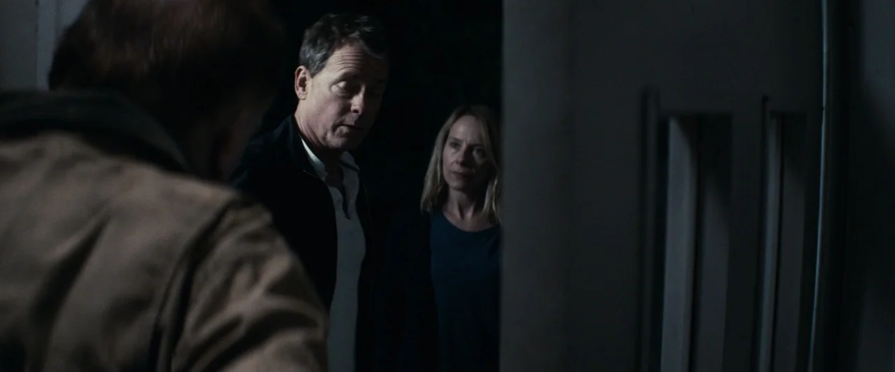 Strange but True (2019) movie still: medium — A dimly lit entryway with dark wall surfaces framing a person in the foreground. A man and a woman…