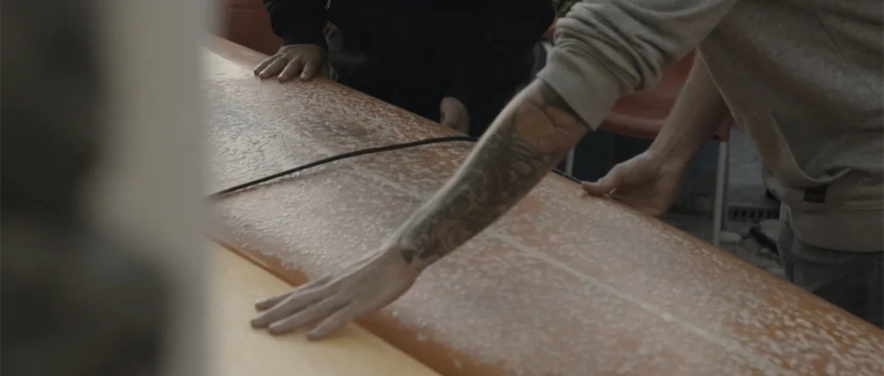 Taking Back the Waves (2017) movie still: medium — A workshop interior focuses on a light brown surfboard being waxed by two people. The subjects…