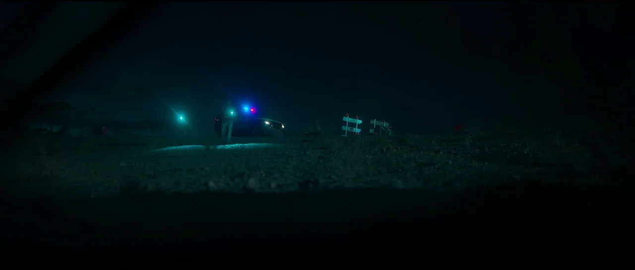 Sympathy For The Devil (2023) movie still: wide — A dark, gravel-covered expanse is illuminated by the flashing lights of a police cruiser. Two…