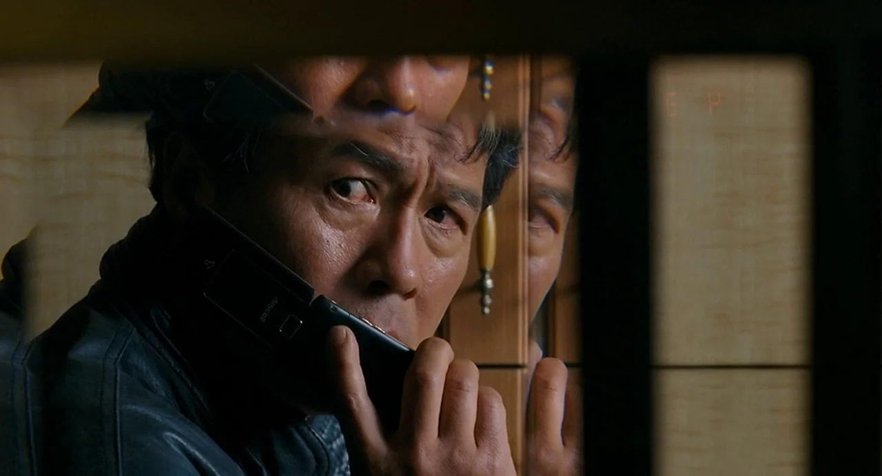 I Saw The Devil (2010) movie still: closeup — A man looks into a mirrored elevator wall while holding a dark flip phone. Multiple reflections of his…