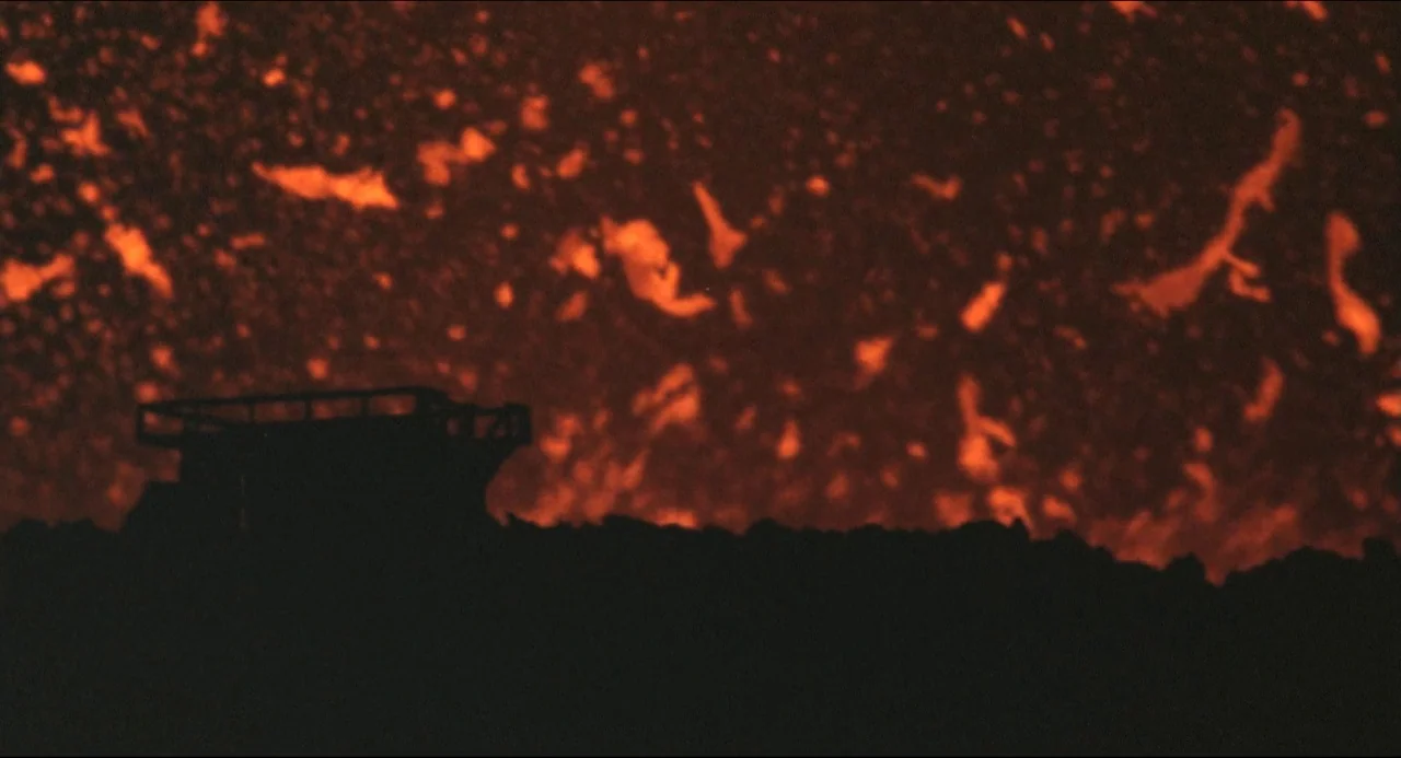 Godzilla vs. Biollante (1989) movie still: wide — An uneven landscape of dark volcanic rock is illuminated by a churning lava flow. A small metallic…