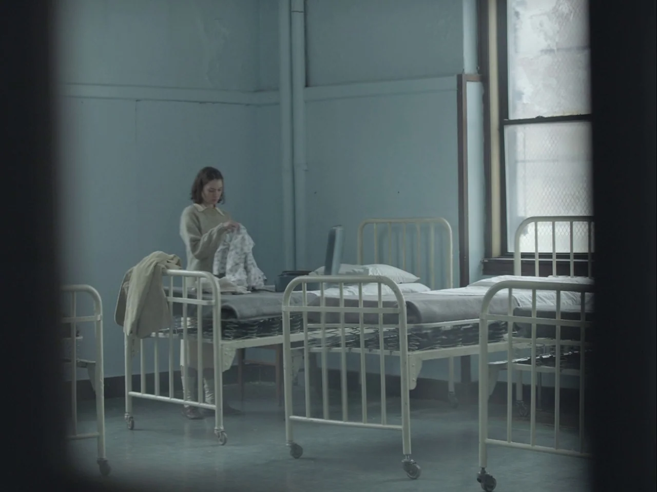 The Mountain (2018) movie still: wide — A sparsely furnished hospital ward features multiple white metal beds with grey blankets. A woman stands by…