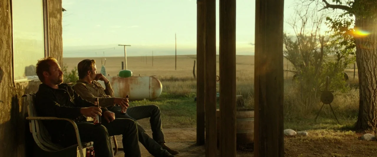 Hell or High Water (2016) movie still: wide — A rural wooden porch overlooks a flat, dry grassy plain during golden hour. Two men sit side by side…