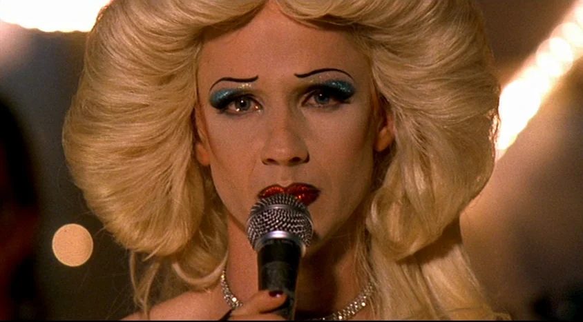 Hedwig and the Angry Inch (2001) movie still: closeup — A performer stands on a dimly lit stage during a musical performance. The subject holds a…