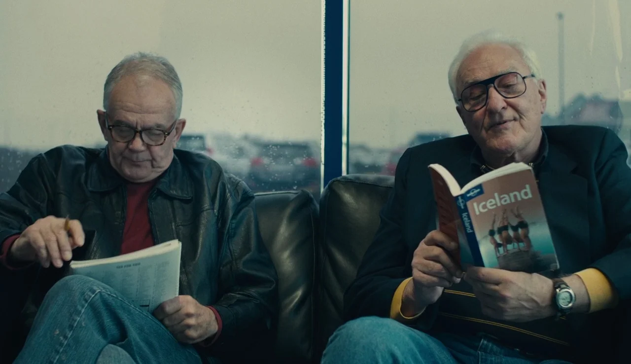 Land Ho! (2014) movie still: medium — Two older men sit on a leather couch in front of a rainy window. One man reads a travel guide about Iceland…