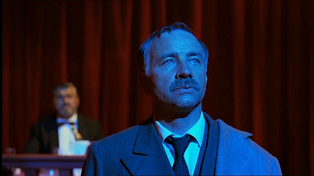 Lola (1981) movie still: medium — A dim room features dark red velvet curtains in the background and a wooden railing. A man with a mustache stands…