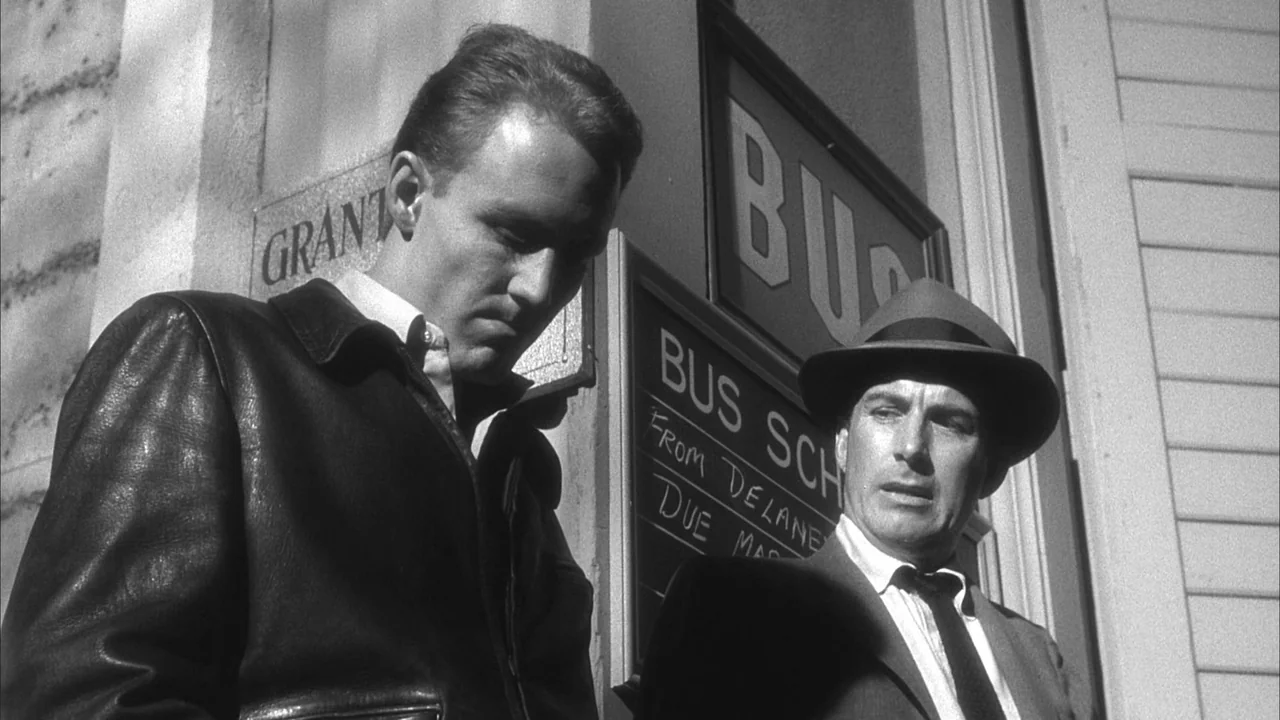 The Naked Kiss (1964) movie still: medium — Two men stand together at an outdoor bus station with a posted schedule board. One man wearing a leather…