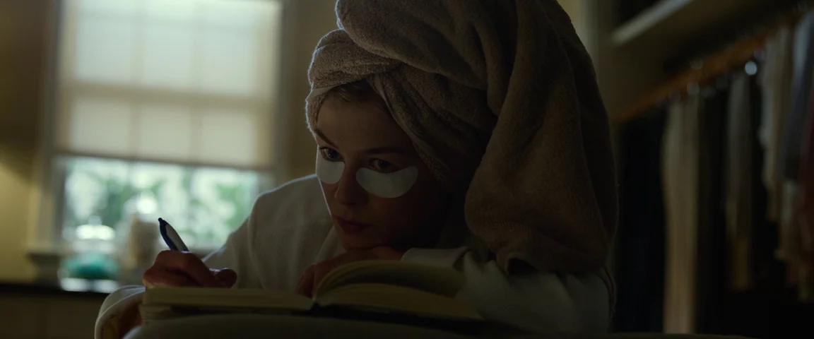 Gone Girl (2014) movie still: closeup — A dark walk-in closet with a soft-lit window visible in the background. A woman wearing a hair towel and…