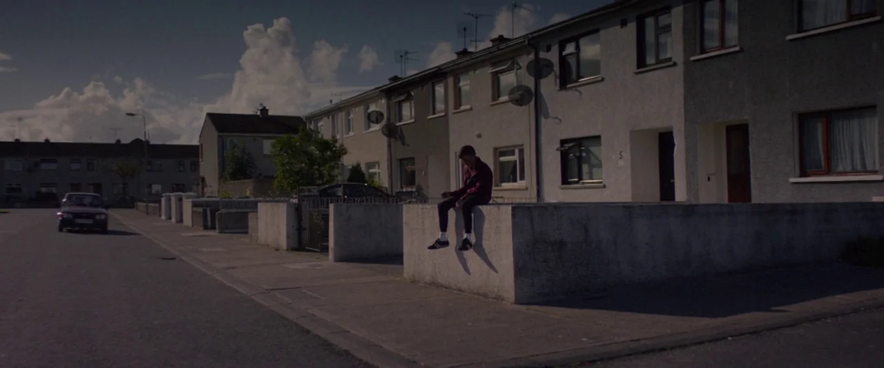 Kissing Candice (2017) movie still: wide — A suburban residential street with row houses and a concrete boundary wall. A lone teenager sits on the…