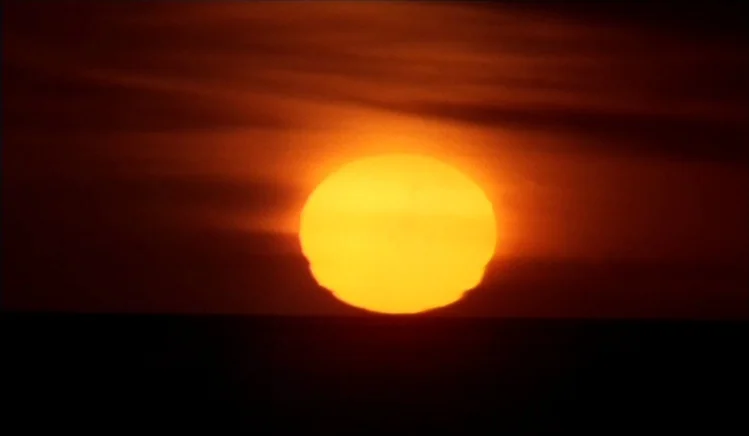 Walkabout (1971) movie still: establishing — A bright yellow sun sets against a dark, layered orange and black horizon. The sun hangs just above the…