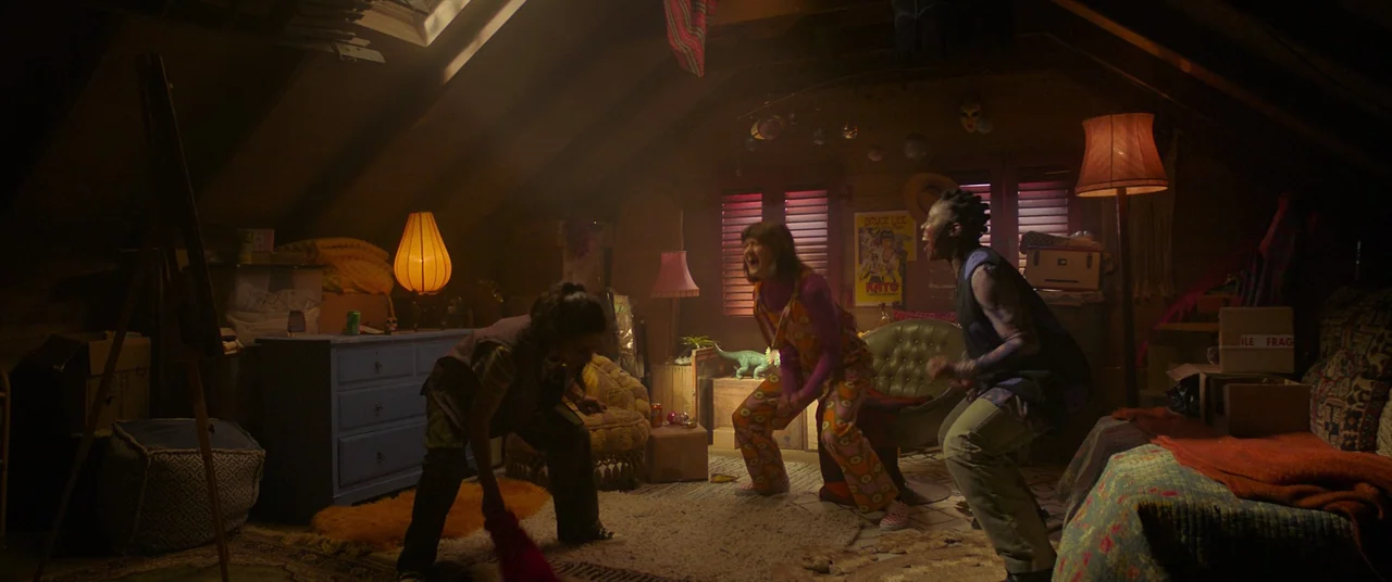 Polite Society (2023) movie still: wide — A cluttered attic space features slanted ceilings, patterned rugs, and assorted vintage furniture. Three…