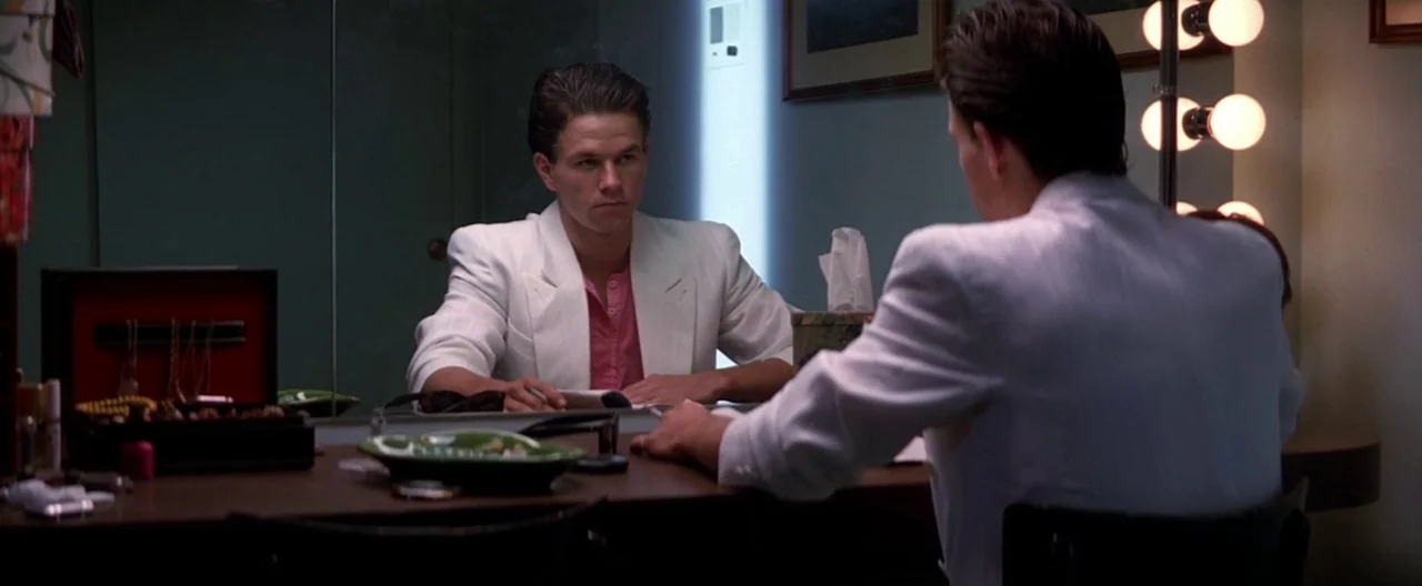 Boogie Nights movie still: medium — A dressing room features a large vanity mirror with exposed incandescent bulbs. A man in a white blazer sits at…