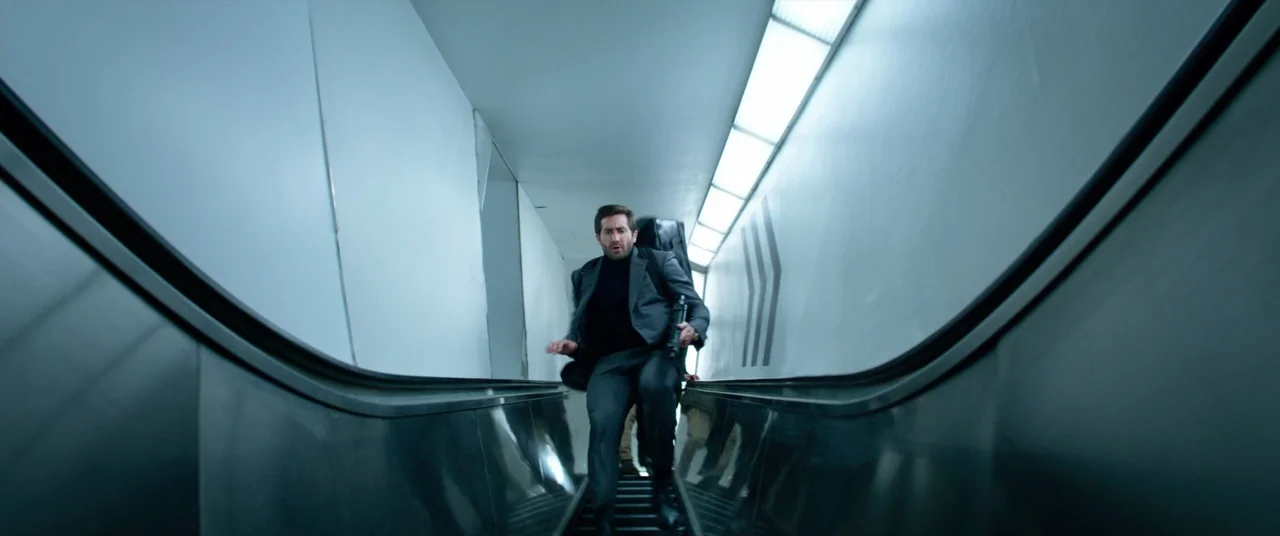 Ambulance (2022) movie still: fullbody — An escalator inside a modern transit space with stark white walls and geometric accents. A man in a suit…