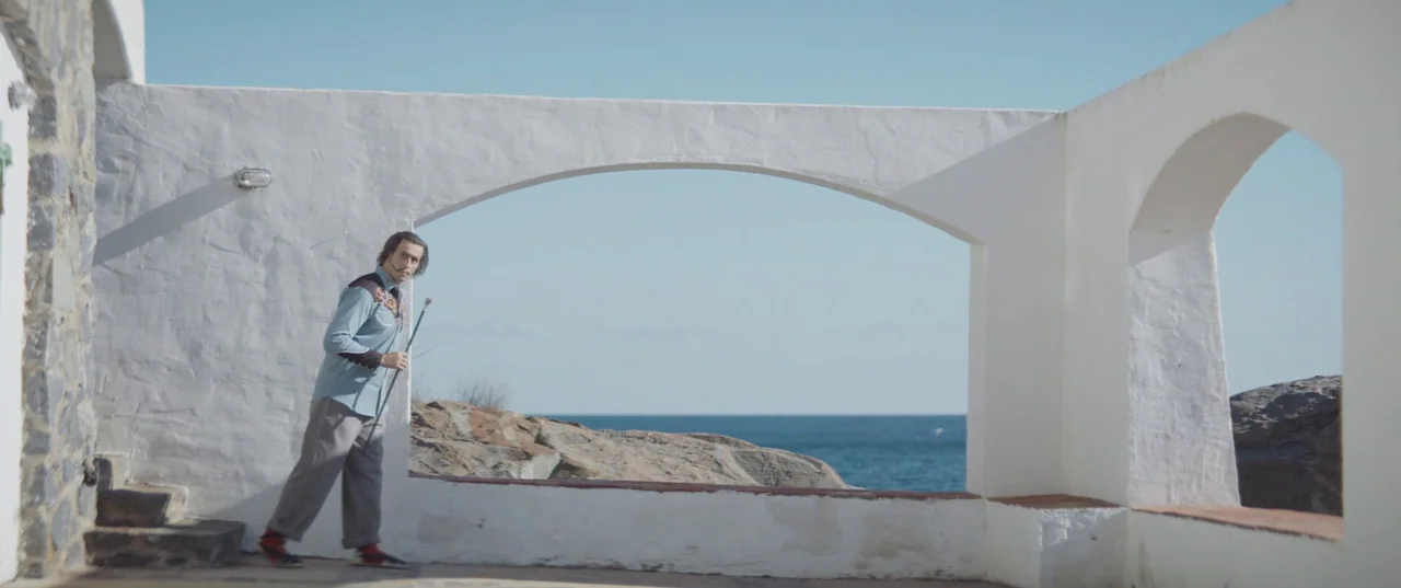 Daaaaaalí! (2023) movie still: wide — A white stucco terrace with arched openings overlooks a rocky coastal landscape. A man wearing a button-down…