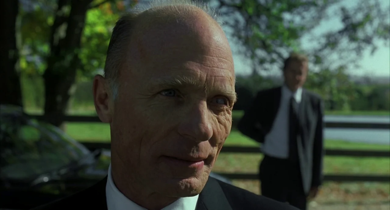 A History of Violence (2005) movie still: closeup — A rural park setting features a wooden fence and trees in the background. An elderly man in a…