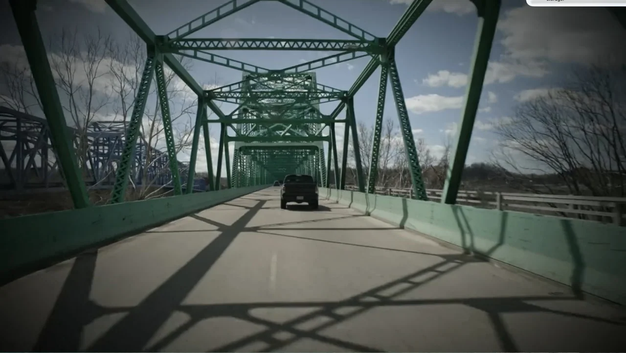 Murder at Carter County Mines (2023) movie still: wide — A long green steel truss bridge extends toward the horizon over a waterway. A black pickup…
