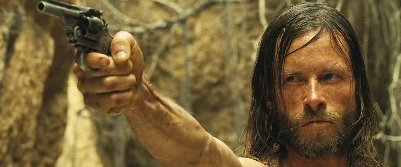 The Proposition (2005) movie still: medium — A man stands in a rugged outdoor canyon with textured earthen walls behind him. He holds a revolver…
