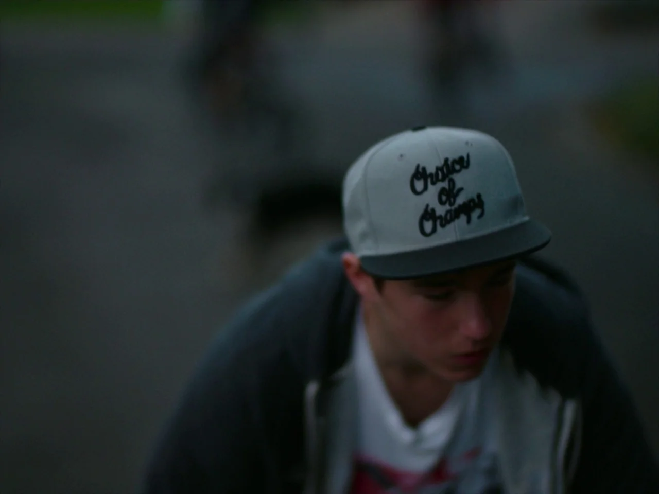 Violet (2014) movie still: closeup — An outdoor paved area serves as the setting for a portrait. A young man wearing a grey baseball cap leans…