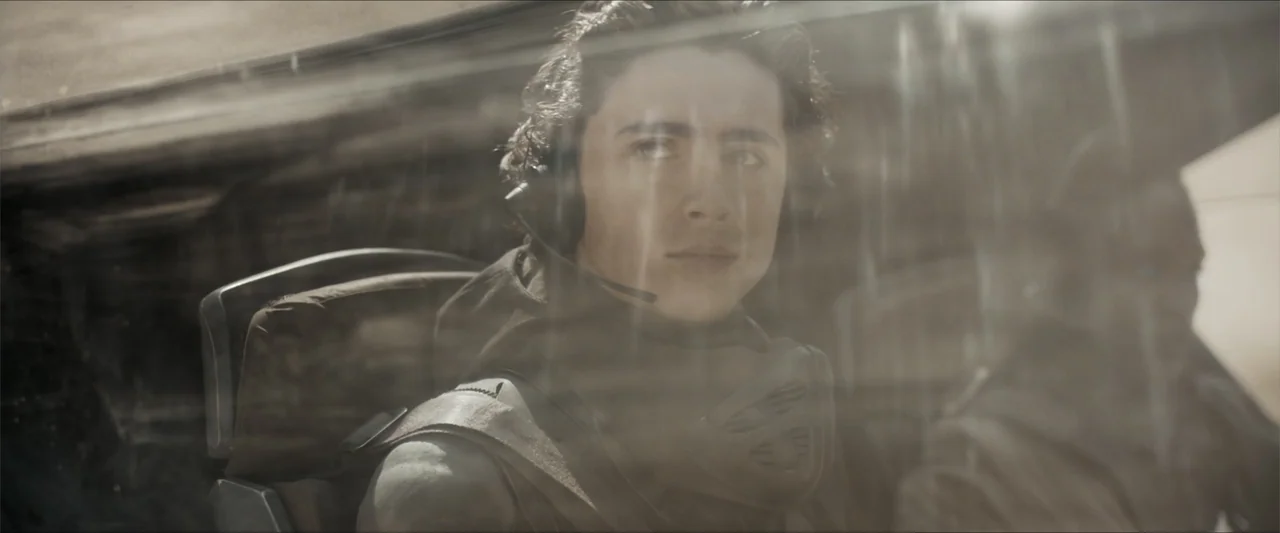 Dune (2021) movie still: medium — A cramped futuristic cockpit interior with diffused light filtering through a thick window pane. Two pilots in…