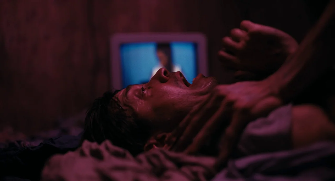 Barbarian (2022) movie still: closeup — A man lies on a bed in a darkened, dimly lit room. A television screen in the background casts a blue glow…